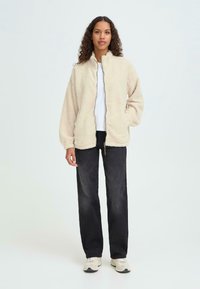 Kreemika värvi fleece jakk kõrge kraega, lukuga ees ja küljetaskudega, koos mustade pükstega ja spordijalatsitega. Sile tekstuur; mugav lõige.