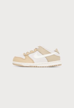 Χαμηλό sneaker με μπεζ και λευκές δερμάτινες επιφάνειες, λευκά κορδόνια, λογότυπο Nike Swoosh σε λευκό χρώμα και μπεζ λαστιχένια σόλα σε ανοιχτό φόντο.