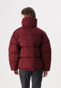 Burgunderfarbene Steppjacke mit hohem Kragen, gestepptem Design und sichtbaren Nähten. Weiche Textur, übergroße Silhouette und elastische Bündchen.