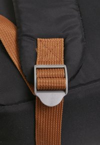 Un sac à dos en tissu noir avec une sangle en toile marron et une boucle en plastique grise. Le gros plan montre le détail de la couture sur la sangle.