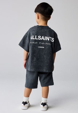 Barn stående med ryggen til, iført mørk oversized AllSaints T-shirt og shorts, hvide sokker og hvide sneakers mod ensfarvet baggrund.