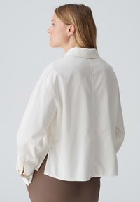 White cotton shirt with a collar, relaxed fit, side slits, and buttoned cuffs. Smooth texture with a minimalistic design.