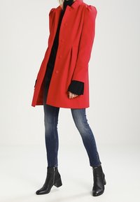 Abrigo de mezcla de lana roja con hombros abullonados, cierres de botones y bolsillos laterales, combinado con jeans oscuros y botas de tobillo.
