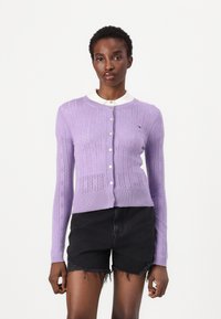 Cardigan de punto lila con un patrón texturizado, dobladillo acanalado, botones frontales y una camisa blanca con cuello debajo. Se lleva con pantalones cortos de mezclilla oscuros.