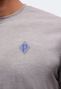 Γκρι βαμβακερό t-shirt με μικρό μπλε λογότυπο σε σχήμα διαμαντιού με το γράμμα "p" στο πάνω μέρος του στήθους κοντά στη λαιμόκοψη.