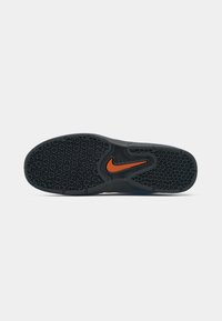 Černá gumová podrážka boty s texturovaným vzorem, obsahující oranžové logo Nike a výrazný design, vhodná pro trakci a odolnost.