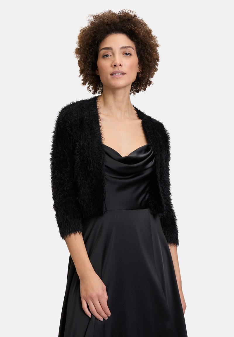 Schwarze Kunstpelz-Bolerojacke mit Dreiviertelärmeln, getragen über einem schwarzen Satinkleid mit einem Wasserfall-Ausschnitt. Weicher, strukturierter Stoff.