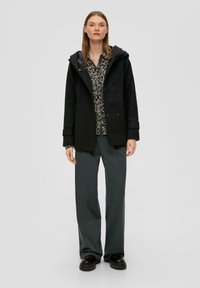 Manteau en mélange de laine noir avec capuche, doté d'une fermeture à snap et de poches latérales. Porté par-dessus un blouse à motif floral et un pantalon large vert foncé.
