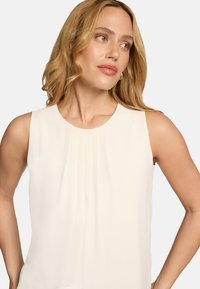 Chemisier sans manches en crème clair, présentant de doux plis au niveau de l'encolure. Tissu lisse et texturé avec un col rond modeste.