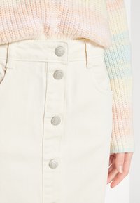 Beige denimskjørt med frontknapper i sølv, to sidelommer og en teksturert strikkegenser i pastellfarger.