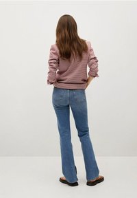 Vaaleanpunainen teksturoitu pusero, jossa käännetyt hihat, yhdistetty vaaleansinisiin liehuhousuihin ja mustiin sandaaleihin. Neutraali tausta.