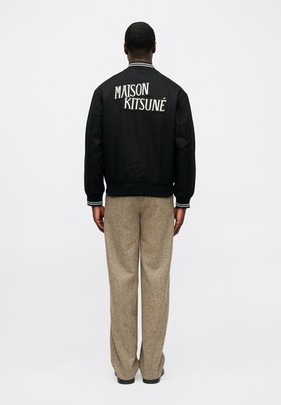 Crna bomber jakna s izvezenim "MAISON KITSUNÉ" na leđima, uparena sa svijetlosmeškim, teksturiranim širokim hlačama.