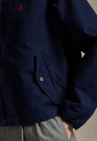 Jachetă ușoară bleumarin din amestec de bumbac, cu închidere cu nasture maro pe buzunar și manchete elastice, având un detaliu brodat cu logo.