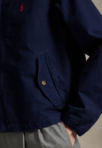 Tumši zila viegla jaka no kokvilnas maisījuma ar brūnu pogu aizdari uz kabatas un elastīgiem manžetēm, ar izšūtu logotipu.