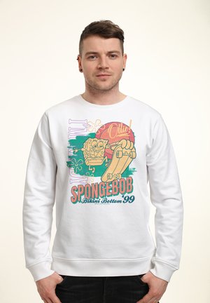 Mand iført hvid sweatshirt med SpongeBob på skateboard og teksten "SPONGEBOB Bikini Bottom '99" på en ensfarvet baggrund.
