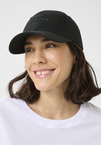 Gorra de algodón negra con visera curva, con un diseño de texto en relieve en la parte frontal. Acompañada de un sudadera blanca con cuello redondo.