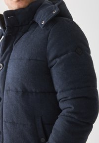 Námornícky modrá vyhrievaná bunda s vysokým golierom, štruktúrovaná tkanina, s gombíkmi a patentkami, so štítkom loga na pleci.