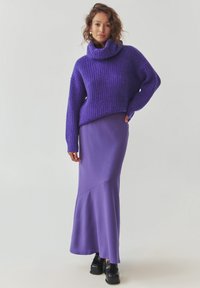 Yli suuri violetti neulepaita, jossa on korkeakaulus, yhdistettynä sileään, pitkään violettiin hameeseen, jossa on vinosauma. Mustat korkokattoiset kengät.