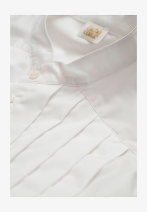Chemise blanche avec un devant plissé, texture lisse, fermeture à boutons et un petit col. Étiquette visible près du col.
