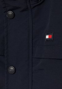 Granatowa kurtka wykonana z textured materiału, z czarnym guzikiem z wytłoczonym napisem "TOMMY" oraz małą łatą z logo w czerwono-biało-niebieskich kolorach.