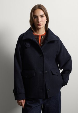 Woman with straight brown hair wearing a dark navy wool coat over an orange and brown buttoned top against a plain light background.