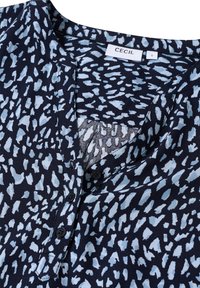 Blusa azul marino con un patrón abstracto en azul claro, cuello redondo, pequeña tapeta de botones y mangas cortas, hecha de un tejido suave.