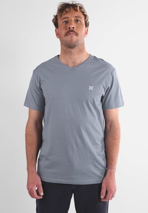 T-shirt gris à col en V en tissu doux. Manches courtes. Présente un petit logo blanc sur la poitrine. Texture lisse, design simple.
