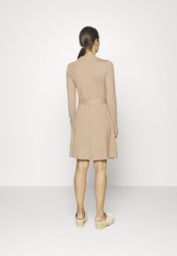 Robe longue beige à manches longues avec un corsage ajusté, une jupe évasée et un détail noué à l'arrière. Portée avec des sabots en bois clair et une texture lisse.
