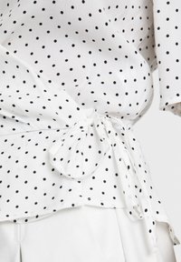 Primo piano di una blusa bianca con pois neri, dotata di una cintura di tessuto annodata intorno alla vita.