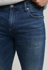 Jeans de mezclilla azul oscuro con un diseño clásico de cinco bolsillos, tela texturizada y costuras visibles a lo largo de las costuras.
