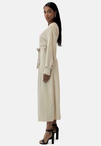 Robe beige longueur cheville avec manches longues et détail de taille nouée. Tissu texturé à finition lisse. Portée avec des sandales à talons hauts noires.