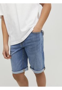 Pantalones cortos de mezclilla en azul claro con dobladillos enrollados y costuras amarillas. Combinados con una camiseta blanca, mostrando una mano en el bolsillo.