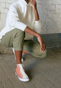 Bílá mikina s kapucí, olivově zelené zkrácené kalhoty a oranžové vysoké tenisky s bílými šňůrkami. Strukturované materiály a neformální styl.