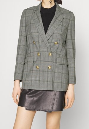 Kobieta w szarej kratkę, dwurzędowej marynarce z złotymi guzikami na czarnym topie i ciemnobrązowej skórzanej mini spódnicy, stojąca na białym tle.