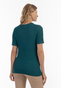 Top tricotat texturat în nuanță de turcoaz, cu un model în formă de scallop, mâneci scurte și un tiv ribbat. Purta cu pantaloni bej, văzut din spate.