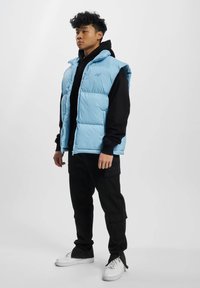 Lyseblå, polstret vest med høj krave, lynlåslukning, båret over en sort hættebluse. Parret med sorte cargo-bukser og hvide sneakers.