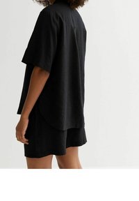 Chemise noire en lin à manches courtes avec un ourlet arrondi, présentant une coupe décontractée et une texture subtile, portée avec un short noir assorti.
