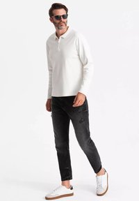 Weißes langärmliges Polohemd mit Reißverschlusskragen, kombiniert mit schwarzen, ausgebleichten Jeans und weißen Sneakers mit braunen Akzenten. Das Model trägt Sonnenbrillen.