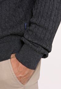 Dunkelgrauer Kabelstrickpullover mit gerippten Bündchen und Saum, aus weichem Material. Hand in der Tasche von hellbeigen Hosen.