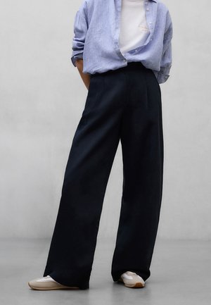 Persoon draagt losse zwarte broek, witte sneakers, wit shirt en een open lichtblauw overhemd met opgerolde mouwen tegen een effen grijze achtergrond.