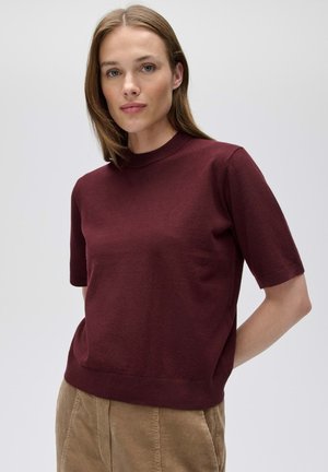 Frau mit glatten hellbraunen Haaren, die einen bordeauxfarbenen Kurzarmpullover und beige Cordhosen vor einem schlichten Hintergrund trägt.