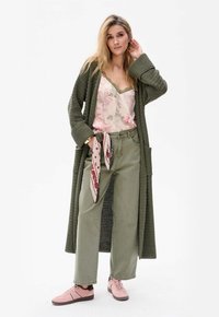 Olijfgroene gebreide cardigan over een bloemen-camisole, gecombineerd met olijfgroene broeken en lichtroze sneakers. Bevat een patroon sjaal rond de taille.