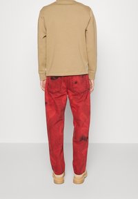 Felpa beige abbinata a jeans rossi leggermente consumati con macchie scure, tenuti su da una cintura beige e stivali chiari.