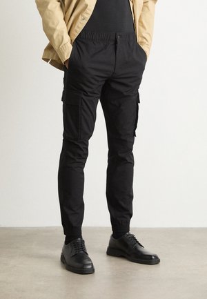 Mann trägt schwarze, schmal geschnittene Cargohose mit elastischen Bündchen, kombiniert mit schwarzen Anzugschuhen und einer beigen Jacke, die Hände in den Taschen.