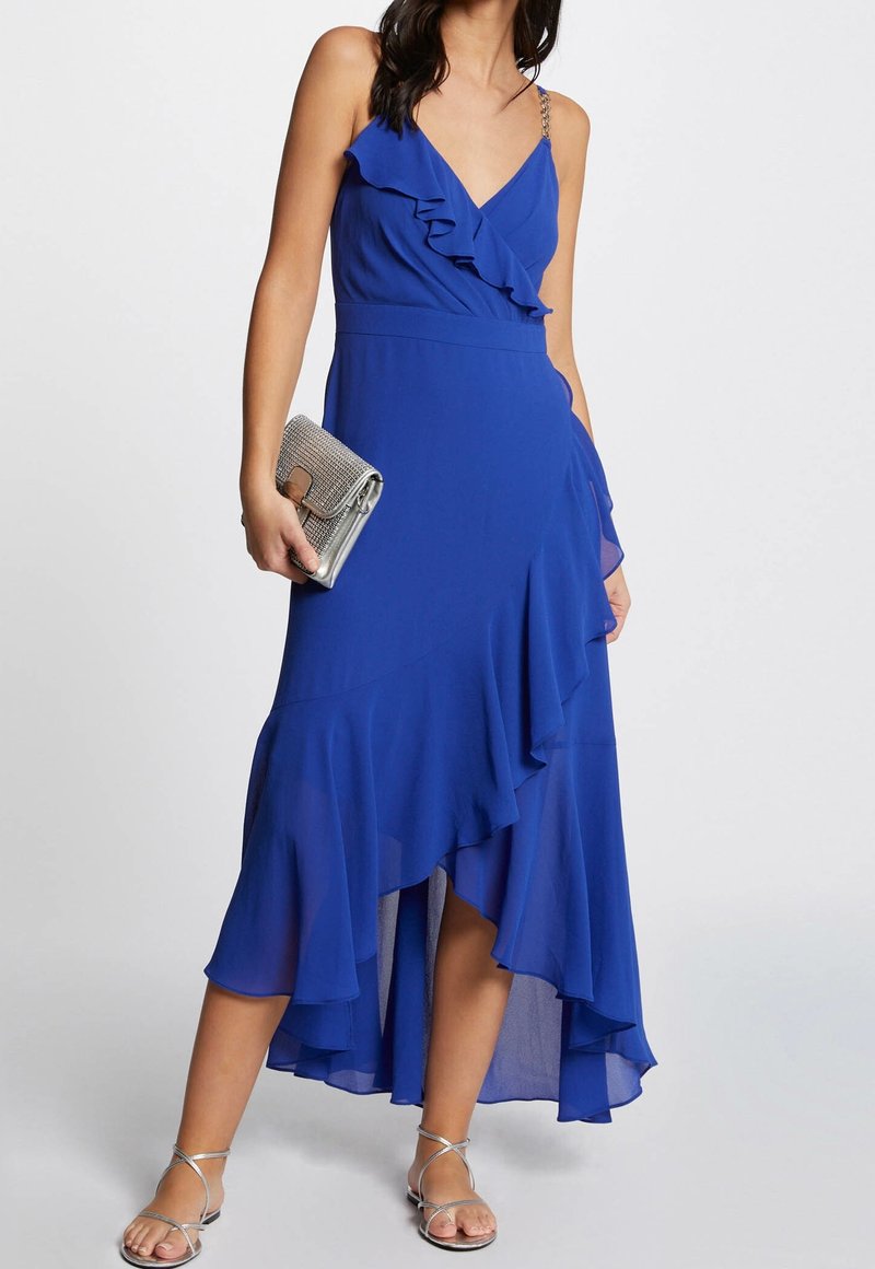 Femme portant une robe bleue sans manches à volants avec des bretelles en chaîne argentée, tenant une pochette argentée et des sandales argentées à brides.
