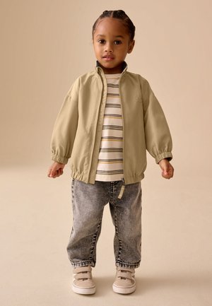 Jeune enfant aux cheveux tressés portant une veste beige zippée, un t-shirt rayé, un jean gris et des baskets beiges, debout sur un fond uni.
