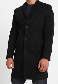 Zwarte wollen jas met een snit kraag, vier knopen aan de voorkant, twee zijzakken en een slanke pasvorm.