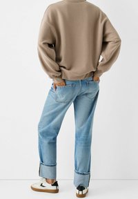 Suéter marrón de cuello redondo, vaqueros de tiro bajo de color azul claro, con puños enrollados y zapatillas blancas con detalles en negro. Tela suave y estilo casual.