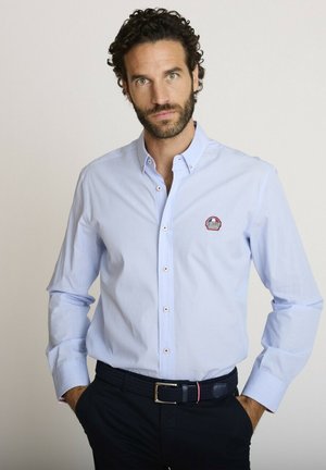 Homme aux cheveux bouclés et à la barbe, portant une chemise bleu clair boutonnée avec un logo et une ceinture noire, mains dans les poches, fond neutre.