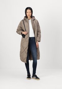 Duga, quilted, smeđa puffer jakna s kapuljačom, koja ima prednji zip i bočne džepove. Nosena preko bijele majice i plavih traperica, u paru s crnim tenisicama.